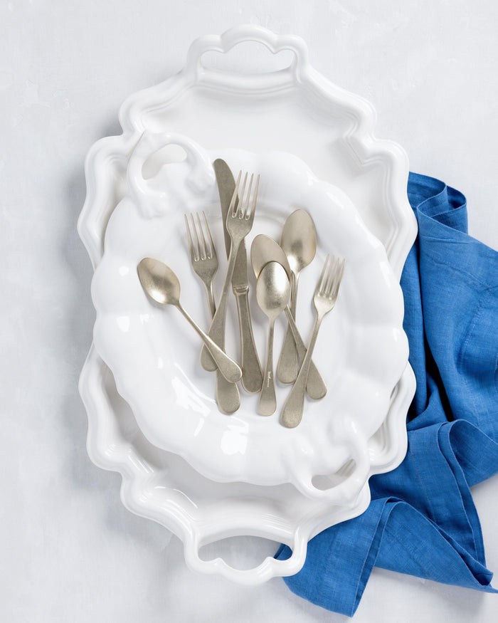 White ceramic oval serving platter with handles, stacked with a smaller platter, silverware, and blue napkin.