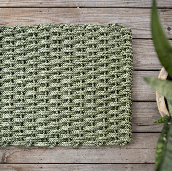 Handwoven Loden Green Rope Doormat on a wooden deck, with a potted plant partially visible.