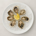 White ceramic serving dish with six oysters on ice and a lemon wedge.
