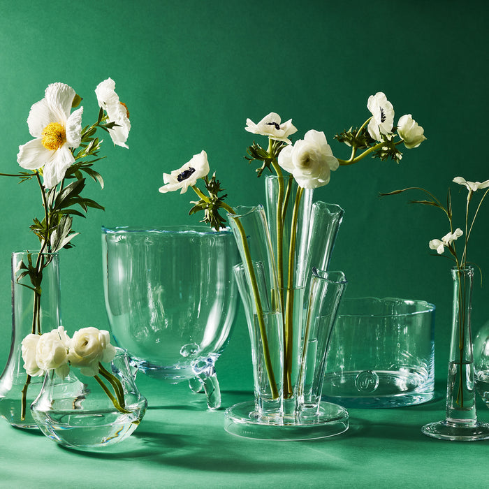 Clear mouth-blown glass Marco Vase with white flowers, surrounded by other clear glass vases on a green background.