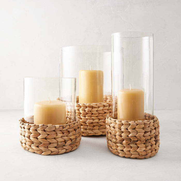 Three glass hurricanes with woven natural hyacinth bases and amber candles.