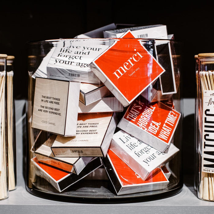 Oversized "Merci" matches in a clear glass jar, featuring red and white boxes with various quotes.
