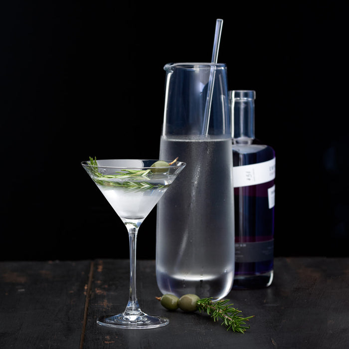 Clear Midtown Martini Glass with a drink, rosemary, and olives, next to a glass carafe and purple bottle.