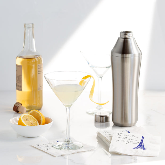 Clear glass Midtown Martini glass with a lemon twist, next to a shaker, bottle, and lemon slices.