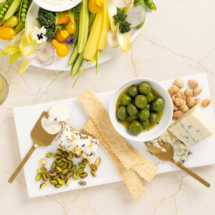 White ceramic rectangular serving platter with olives, cheese, crackers, pistachios, and goat cheese.