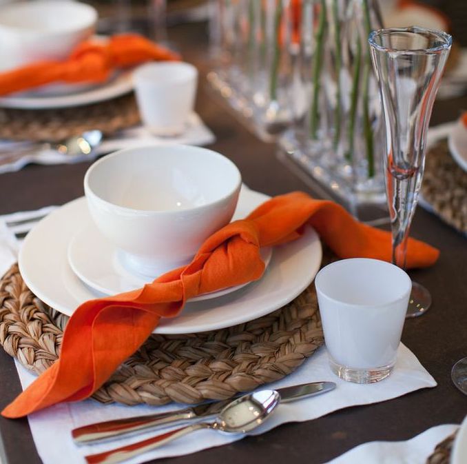 White ceramic footed bowl on a plate with an orange napkin, part of a table setting.