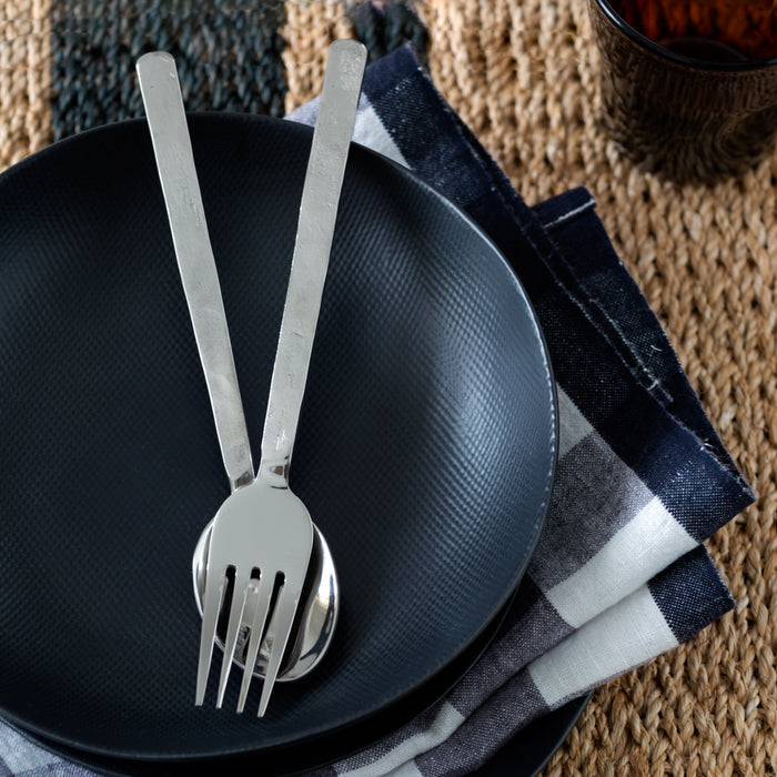 Orleans Stainless 5-piece flatware set with hand-hammered texture on black plates, plaid napkin, and woven mat.