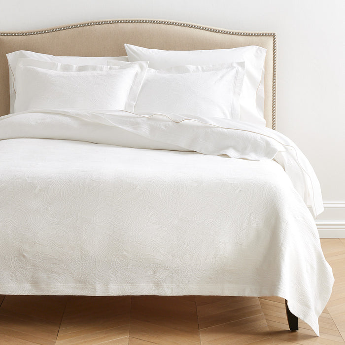 White cotton matelassé shams with subtle paisley pattern on a bed with a beige headboard.