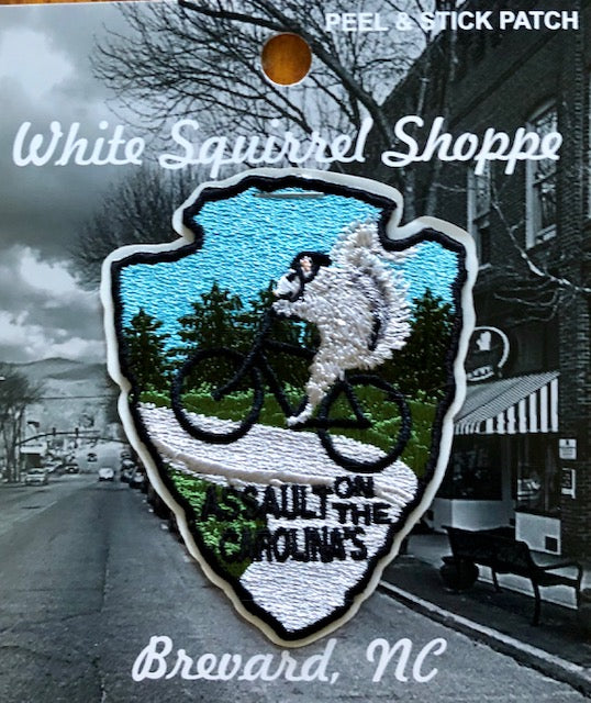 An embroidered peel and stick patch featuring a white squirrel on a mountain bike with the text 'Assault on the Carolinas' and 'Brevard, NC'.
