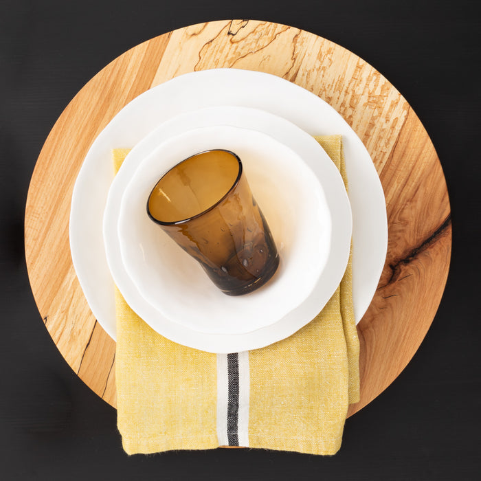 Pebble White ceramic dinner plate, salad plate, and bowl set with a brown glass on a yellow napkin and wood charger.