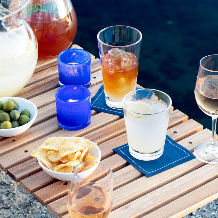 Clear acrylic double old-fashioned glass with a lemon slice, on a blue coaster, part of an outdoor poolside setting.