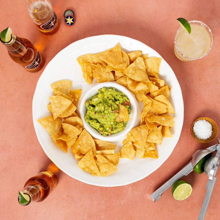 White plate with guacamole and tortilla chips, surrounded by beer bottles, a margarita, salt, and a citrus juicer.