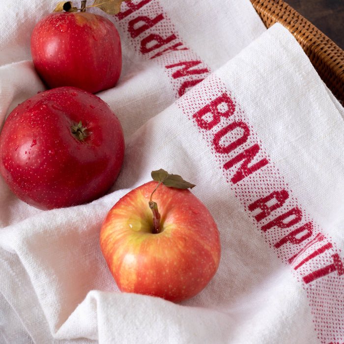 White linen "Bon Appetit" hand towel with red text, folded with three red apples.