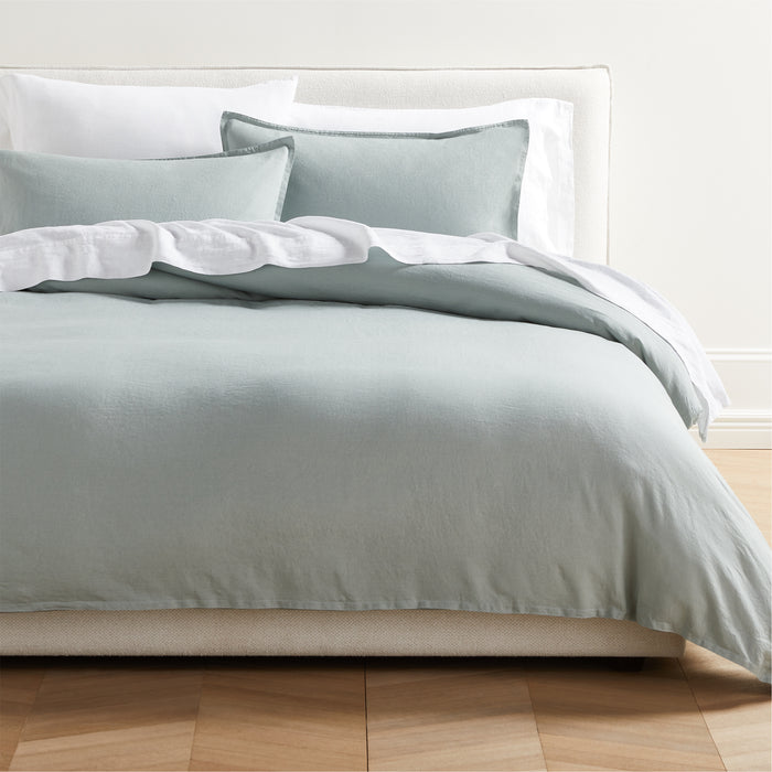 Sage green washed linen pillow shams with a 2" flange and double-needle stitching on a bed.