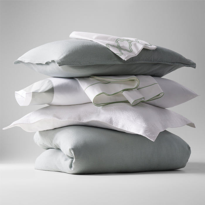 Stack of sage green washed linen pillow shams, white pillows, and embroidered white linens.