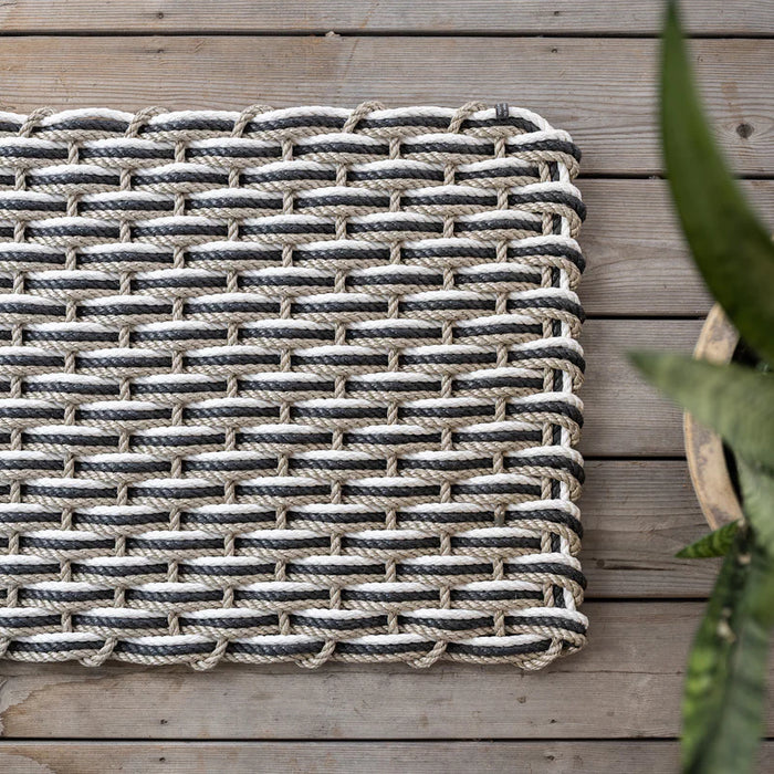 Handwoven sand, charcoal, and pearl rope doormat on a wooden deck, with a plant partially visible.
