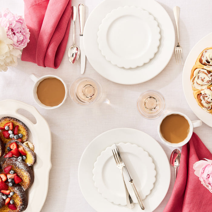 White ceramic scalloped salad plate set on a table with breakfast, pink napkins, and peonies.