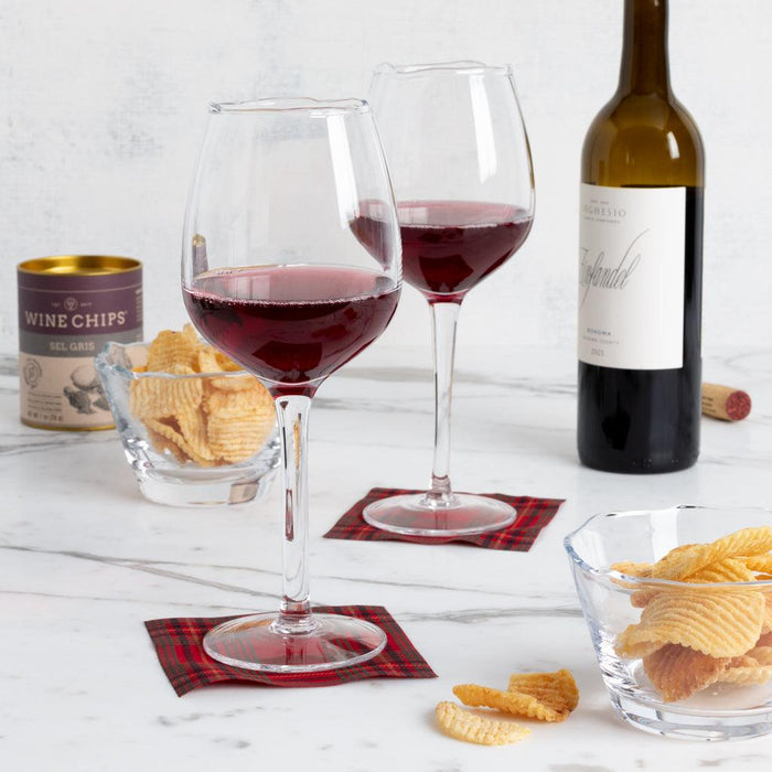Two clear wine glasses with red wine on plaid coasters, a bottle of Zinfandel, and bowls of potato chips.