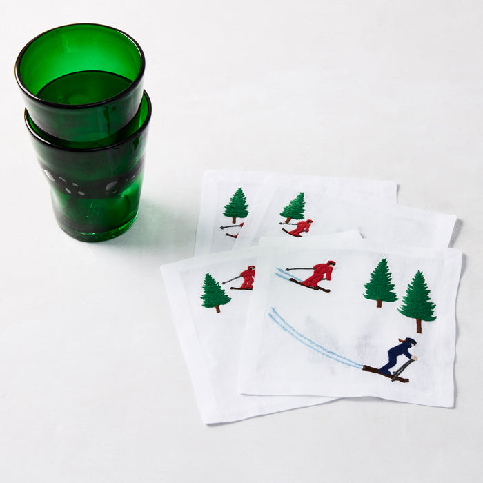 Set of 4 white linen 5.5" square coasters with embroidered skiers and green trees, next to green glasses.
