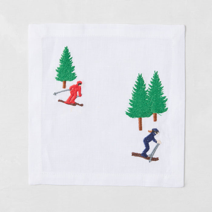 White linen cocktail napkin with embroidered red and blue skiers and green pine trees.