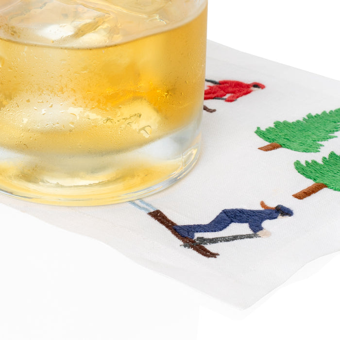White linen coaster with embroidered skiers and green trees, under a glass of iced drink.