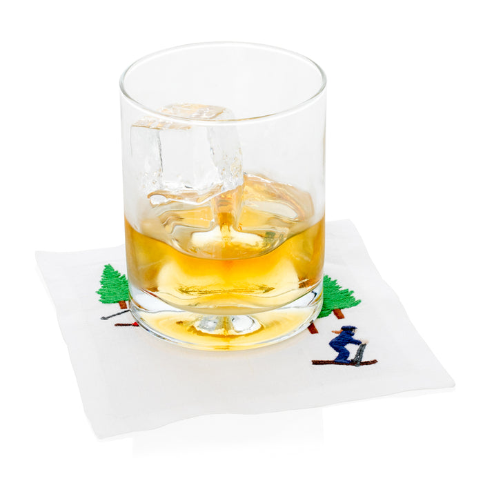 White linen coaster with embroidered skier and green trees, under a glass of amber liquid.