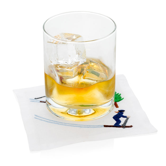 White linen coaster with embroidered blue skier and green tree, under a glass of whiskey with ice.