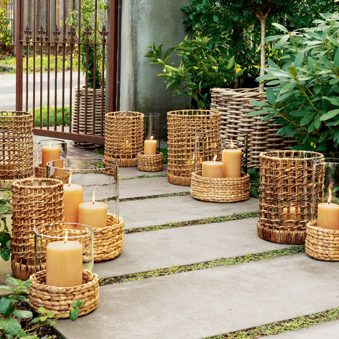 Small glass hurricane with a woven water hyacinth base and amber candle, displayed outdoors.