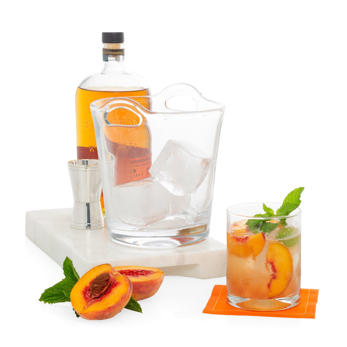 Small clear glass ice bucket with integrated handles, filled with ice cubes, on a white marble tray.