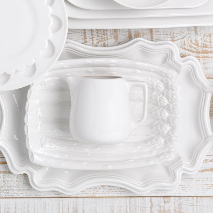 White ceramic milk pitcher on a white rectangular platter with asparagus relief, surrounded by other white serveware.
