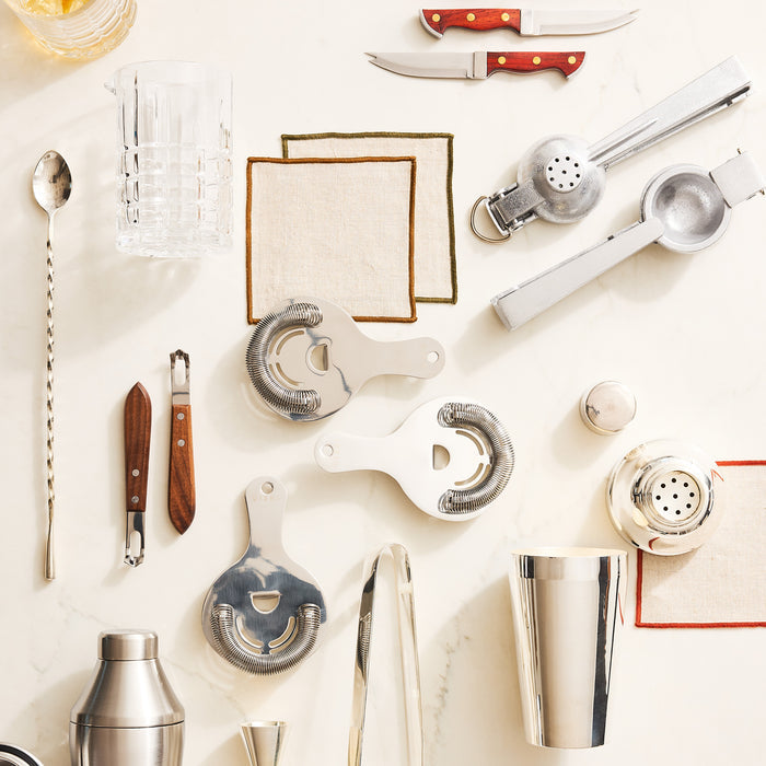 Stainless steel Hawthorne cocktail strainer with spring, shown among other bar tools on a white marble surface.
