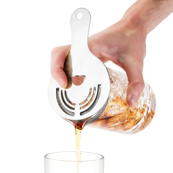 Hand holding a silver stainless steel Hawthorne cocktail strainer, pouring a drink from a glass mixer.