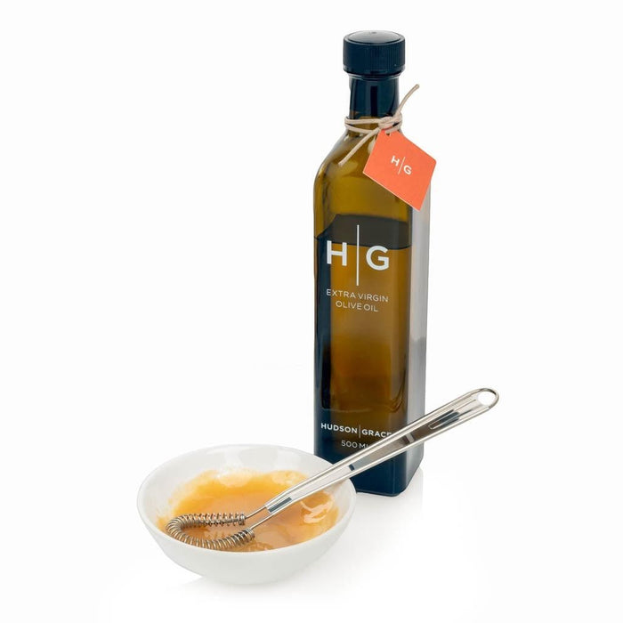 Stainless steel mini whisk in a white bowl with vinaigrette, next to a dark olive oil bottle.