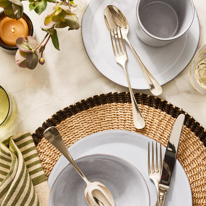 Stone grey terracotta mug, bowl, and plate set with silver flatware on a woven placemat.