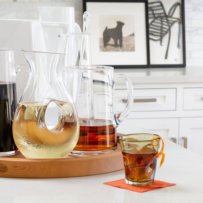 Clear glass tall tapered bar pitcher with handle, holding amber liquid, on a wooden tray.