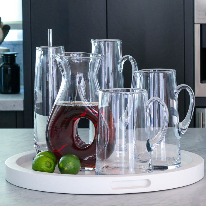 Clear glass tall tapered bar pitcher with handle, filled with iced tea, surrounded by other clear glass pitchers on a white tray.