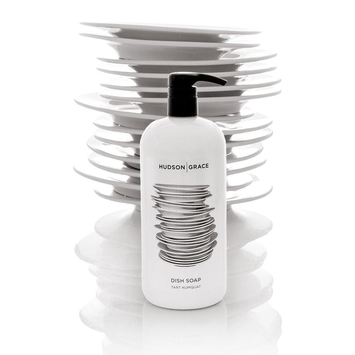 White Hudson Grace Tart Kumquat Dish Soap bottle with black pump, in front of stacked white plates.