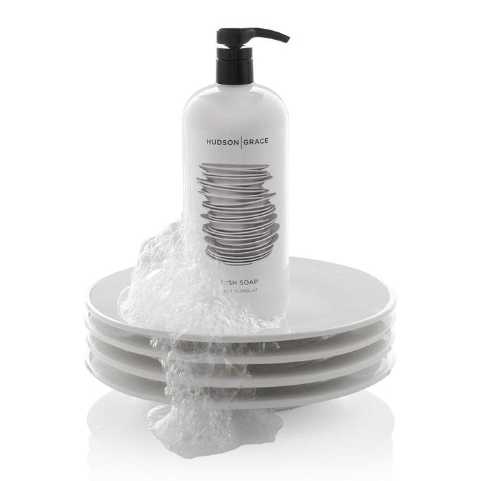 White HG Tart Kumquat Dish Soap bottle with black pump, 32 oz, on a stack of white plates with suds.