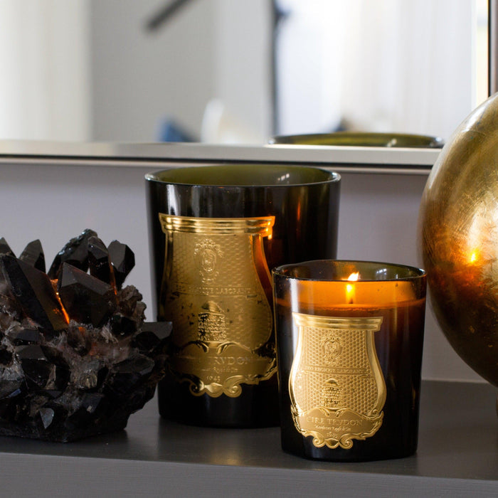 Trudon Spiritus Sancti scented candles in dark glass vessels with gold labels, one lit, next to a black crystal.