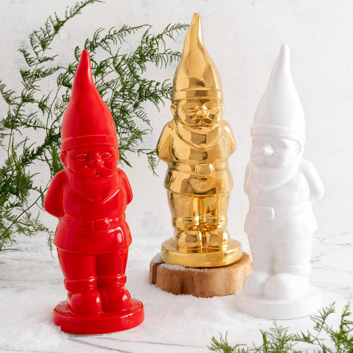 Three ceramic holiday gnomes: red, gold, and white, standing in faux snow with greenery.