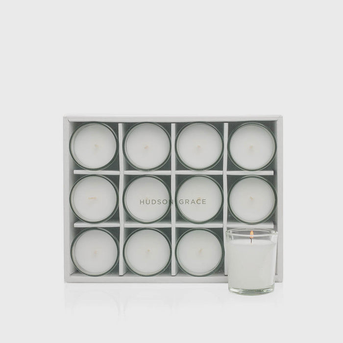 Set of 12 white paraffin votive candles in clear glass cups, one lit, in a white storage box.