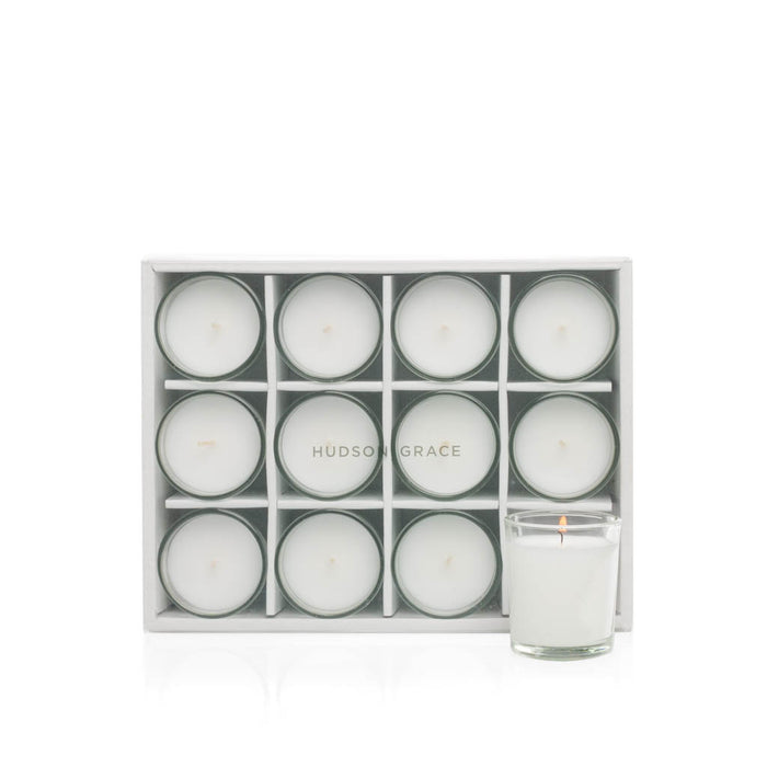 White paraffin votive candles in clear glass cups, set of 12, with one lit candle.