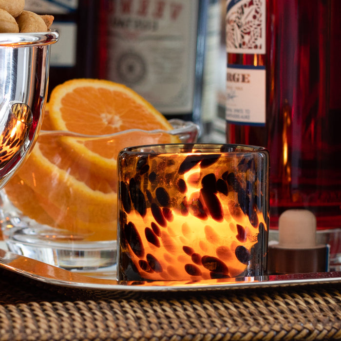 Lit votive candle in a clear glass holder with dark brown leopard spots, next to orange slices and bottles.