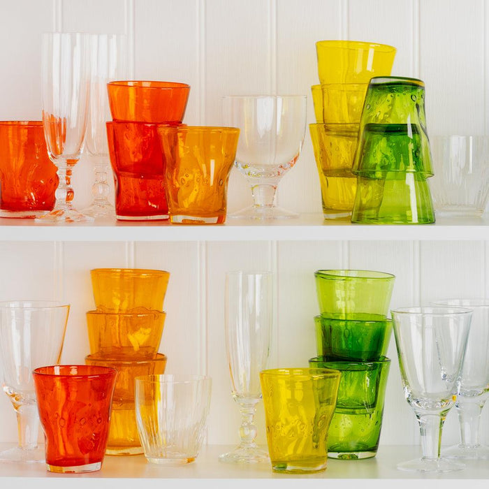 Collection of colorful Henri mouth-blown glassware in yellow, orange, green, and clear on white shelves.